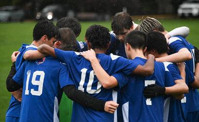 Boys on a soccer team huddled on a field in the rain wearing blue and white jerseys with their heads down and their arms around each others shoulders