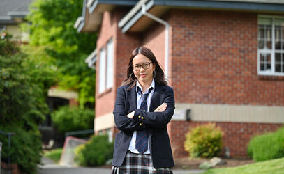A student in the SMUS uniform blazer, tie and tartan skirt stands with arms crossed in front of a brick campus building.