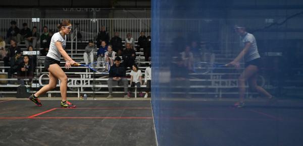 A squash player hits the ball in a glass court, and is reflected in the side wall.