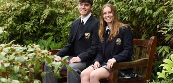 Head Prefects Ryan and Talia pose for a photo while sitting on a wooden bench in front of green flora.