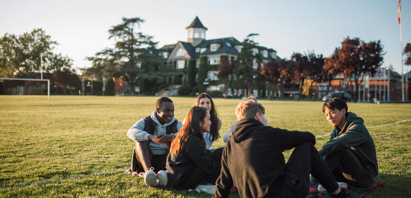 A group of senior students sit together on the field with the school in the background.
