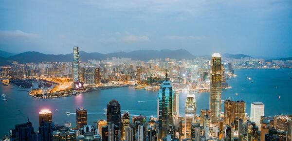 Hong Kong skyline at dusk, with the building lights on and a light fog, overlooking a river.