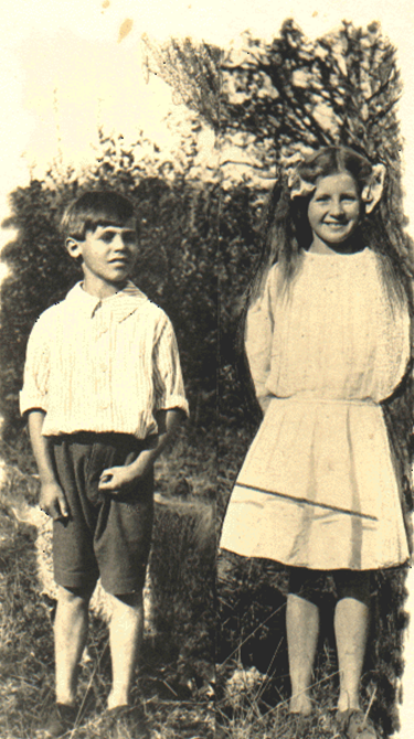 An archival photo of two children, a boy and a girl, standing outside smiling for the camera