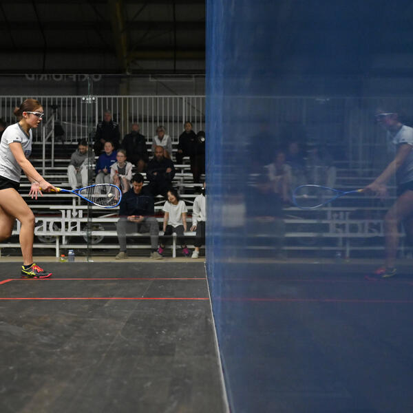 A squash player hits the ball in a glass court, and is reflected in the side wall.