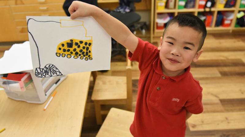 A Kindergarten student proudly holds up a piece of hand-drawn artwork