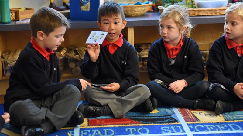 A group of Kindergarten students sit on the carpet while one of them holds up a card with elves on it and speaks.