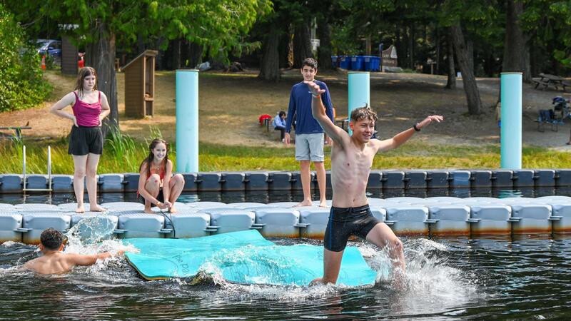 Coed group of campers playing on waterfront with one boy running off the end of a floating dock into the water