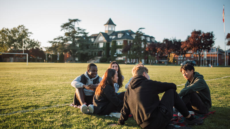 A group of senior students sit together on the field with the school in the background. 