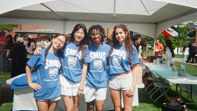 Four students in matching blue SMUS Alumni Weekend T-shirts pose arm in arm under a white tent at an outdoor campus event.