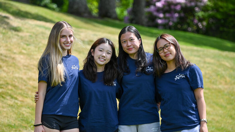 Four students in matching navy Symons House T-shirts smile arm in arm on a grassy slope with flowering rhododendrons behind them.