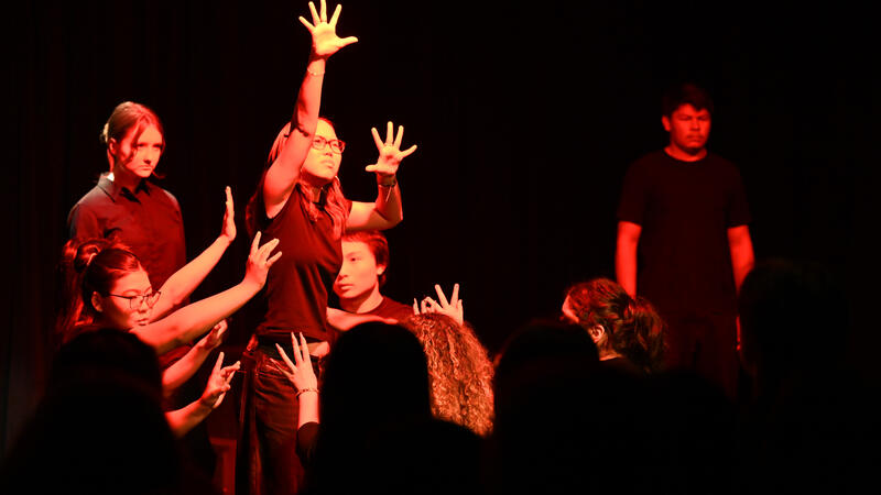 Students dressed in black perform a movement piece under red stage light, one reaching upward as others stretch their hands toward them.