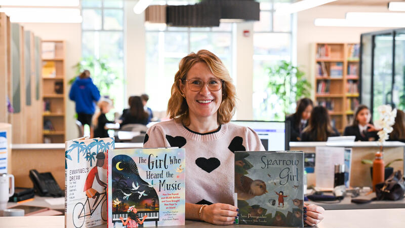 Sarah Bailey, Middle School Librarian, smiles behind a library desk displaying the picture books Emmanuel’s Dream, The Girl Who Heard the Music and Sparrow Girl.
