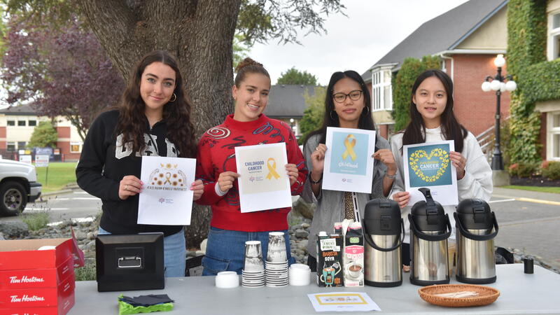 Four students hold childhood cancer awareness posters behind a table of coffee urns, cups and Tim Hortons boxes set up outside on campus.