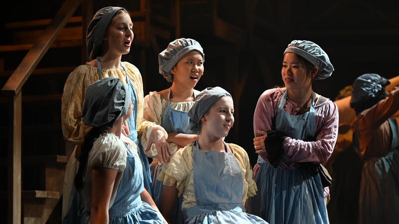 Cast members in bonnets, aprons and period dresses sing together on a dimly lit stage during a school musical.