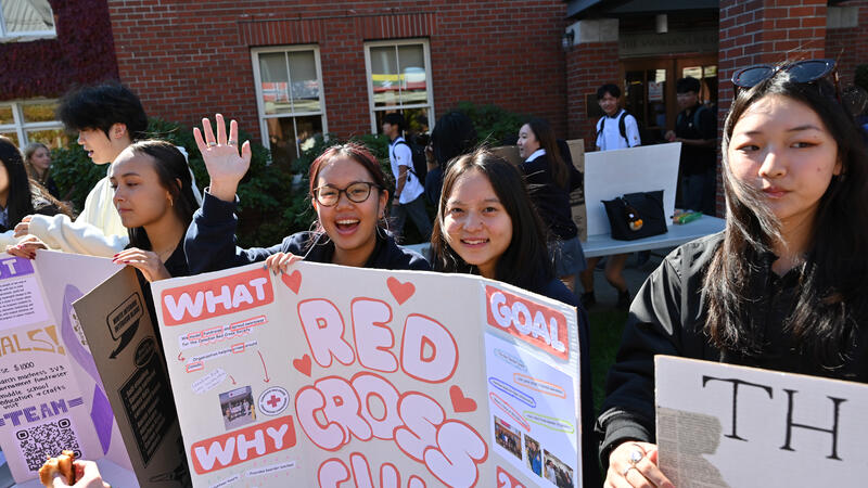 Students hold a hand-decorated Red Cross Club poster board outside the Snowden Library, one waving at the camera.