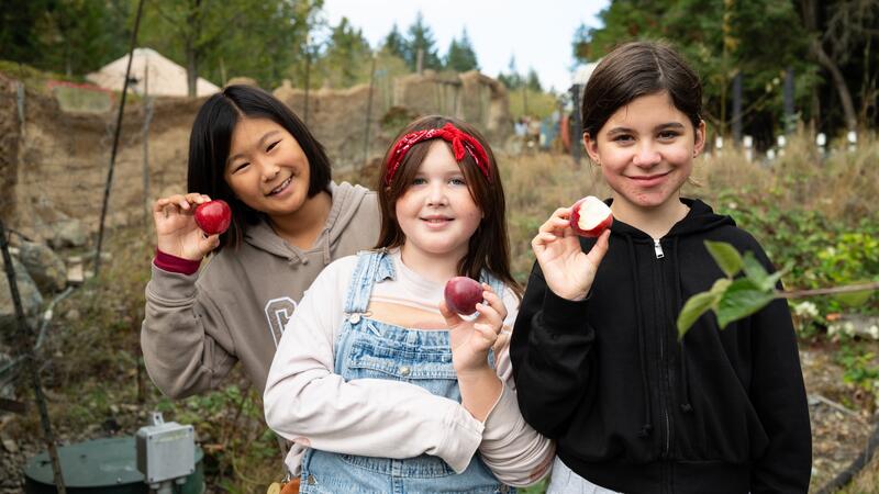 Three students pose with small apples they have picked with the farm trees in the background.