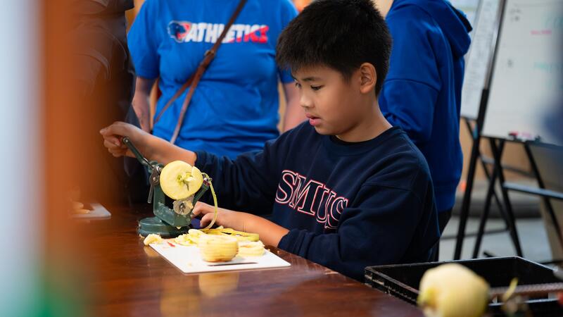 One student uses a mechanical apple peeler to peel a pile of apples. 